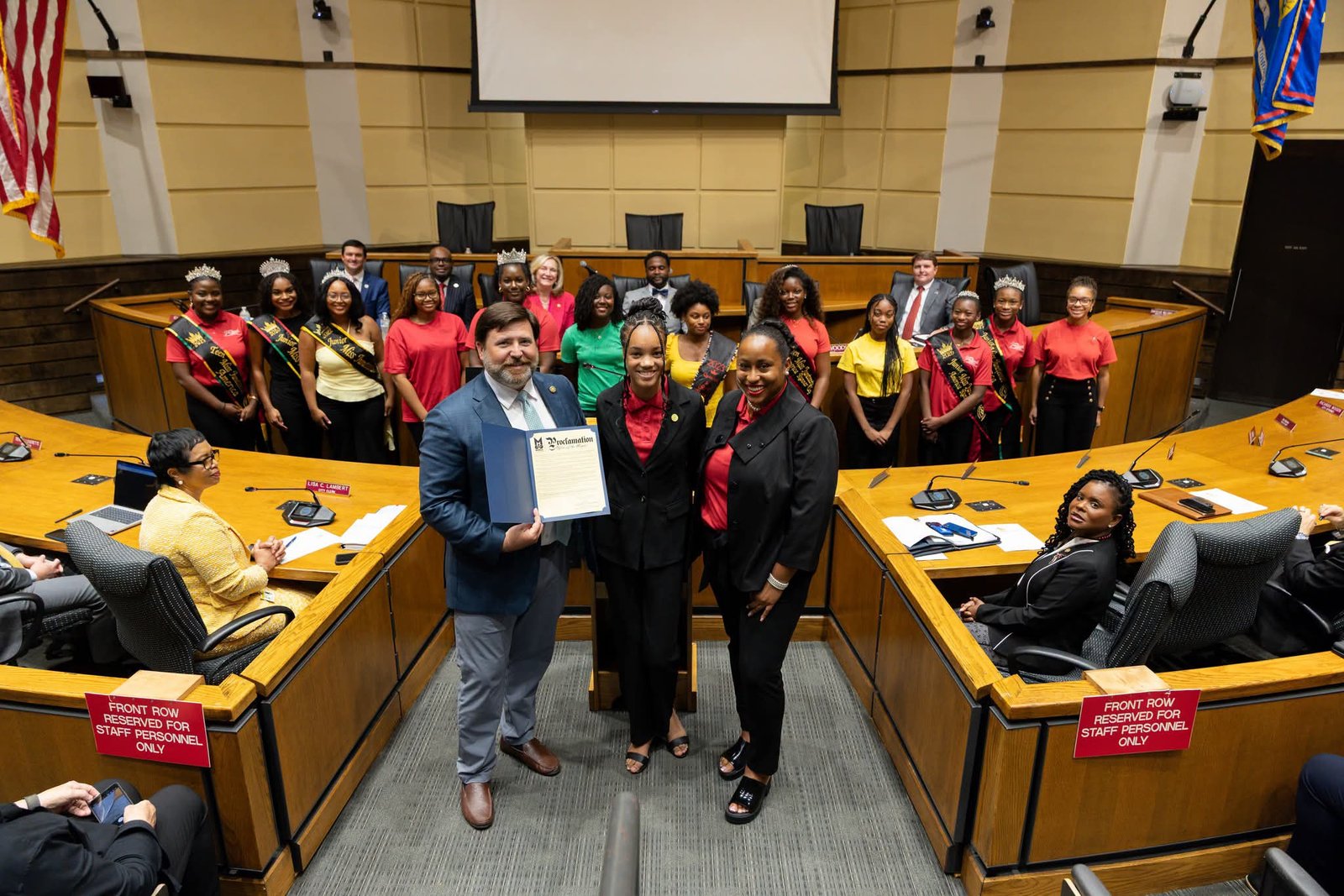 Inside the Mobile City Council chambers, Mayor Spiro Cheriogotis stands in the center foreground holding open an official blue proclamation folder. Two program representatives in formal black business suits stand beside him. In the background row directly behind them, a group of young Miss Juneteenth America delegates stand side-by-side wearing sashes and pageant crowns. 
