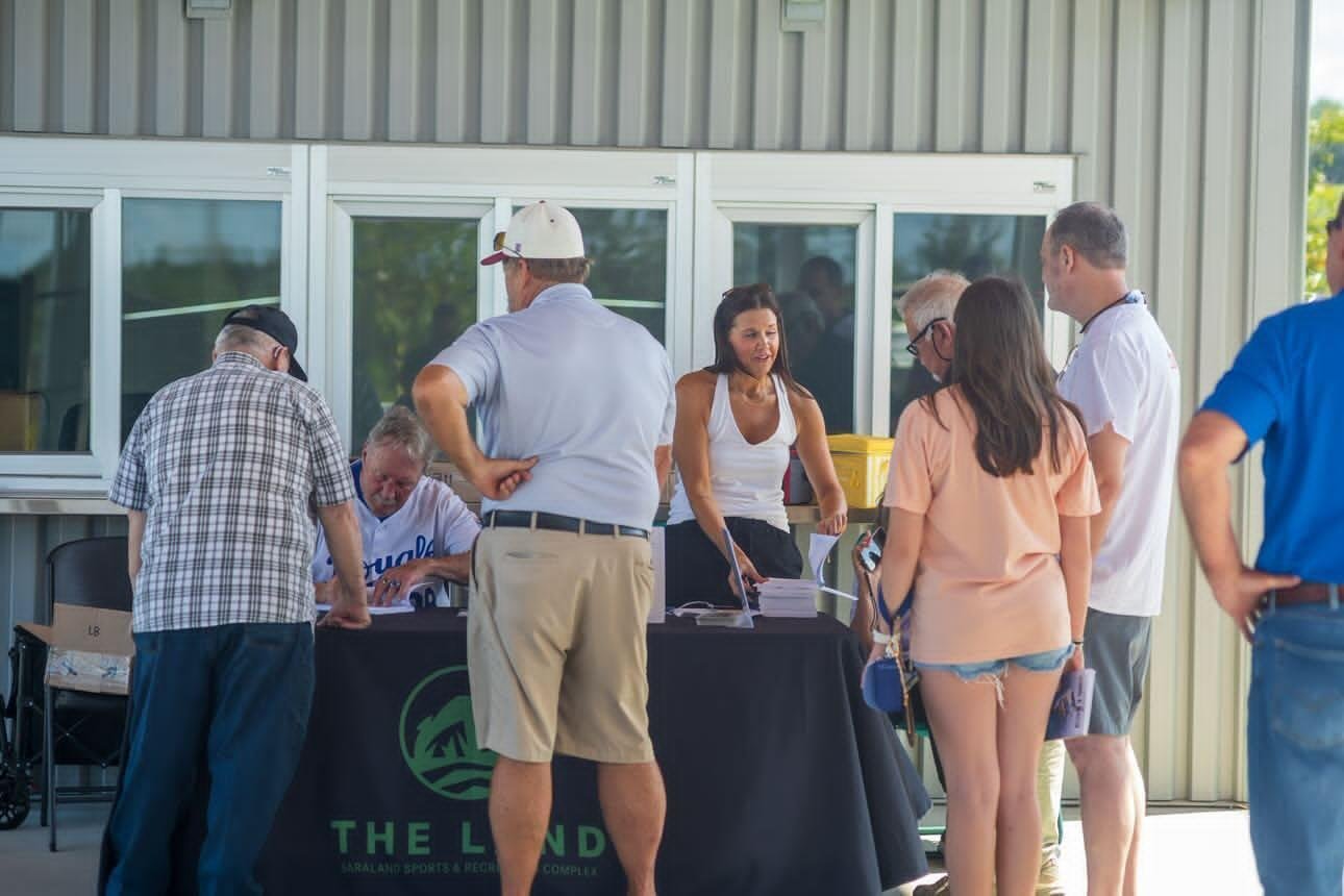 Former MLB player Randy McGilberry sitting at a branded outdoor table for 'The Land' complex, signing autographs and books for a line of community members during his book launch event.