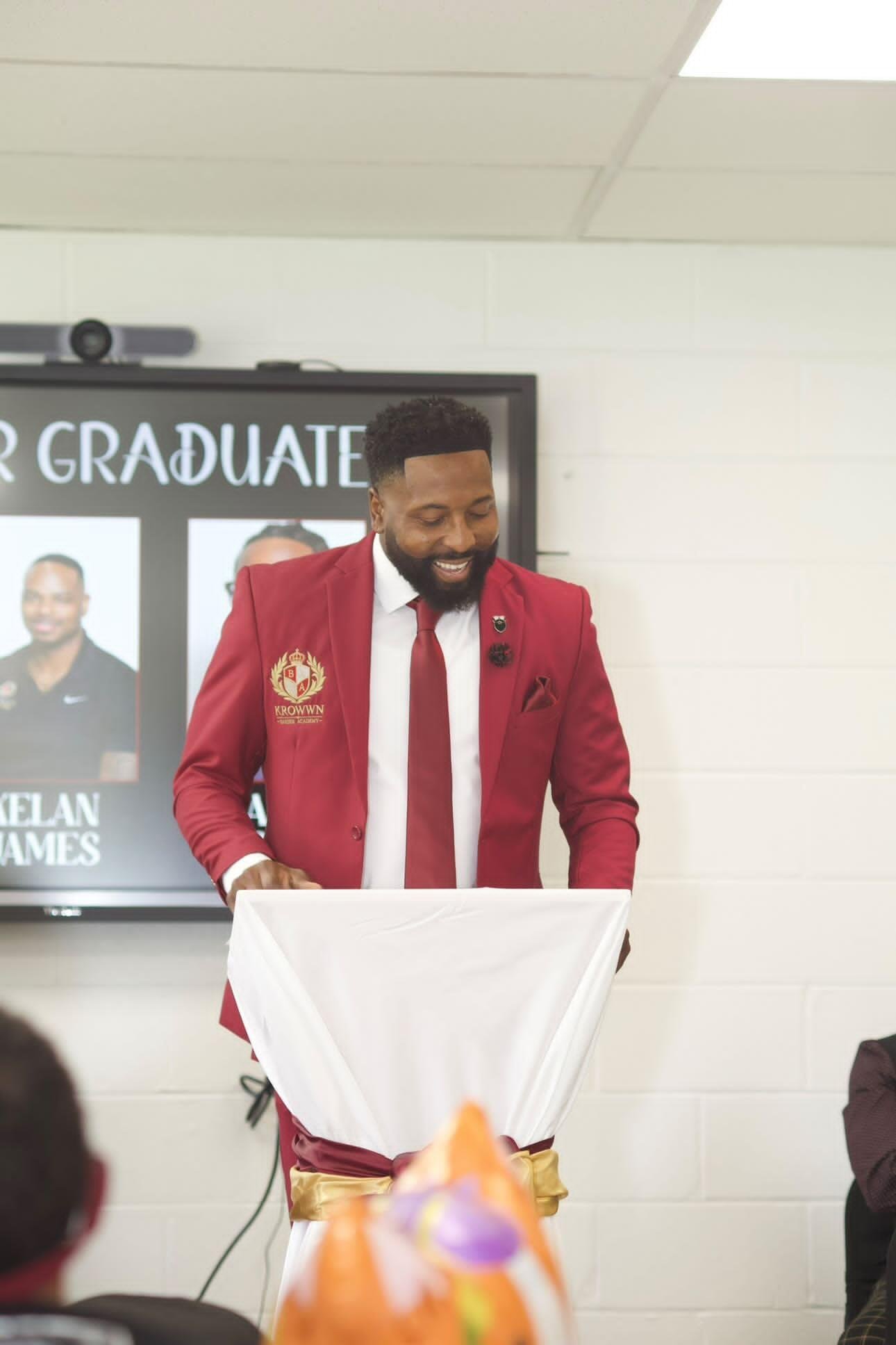 Houston Owens in a branded red suit speaking at the Krowwn Barber Academy inaugural graduation banquet, 2026.