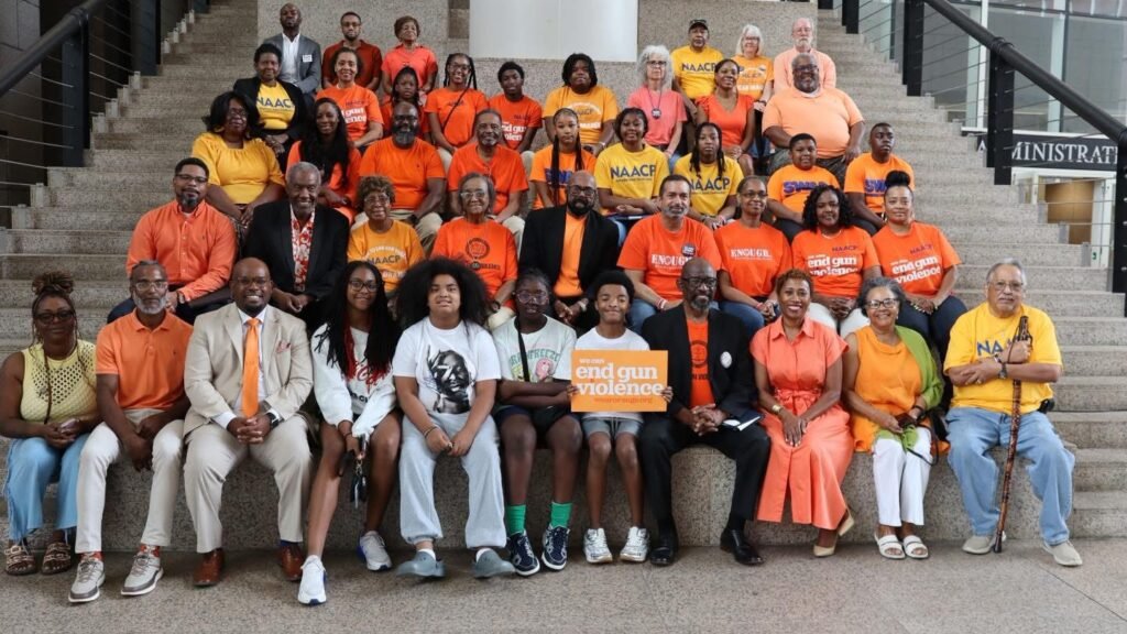 Large group photo of community leaders, residents, youth, and advocates gathered on the steps inside Mobile Government Plaza during a Project Wear Orange event. Participants are dressed primarily in orange shirts and clothing, many displaying messages supporting the effort to end gun violence. Several individuals wear NAACP apparel, while others hold signs promoting violence prevention and community safety. The diverse group poses together in a show of unity, solidarity, and commitment to raising awareness about gun violence and supporting safer communities in Mobile, Alabama.