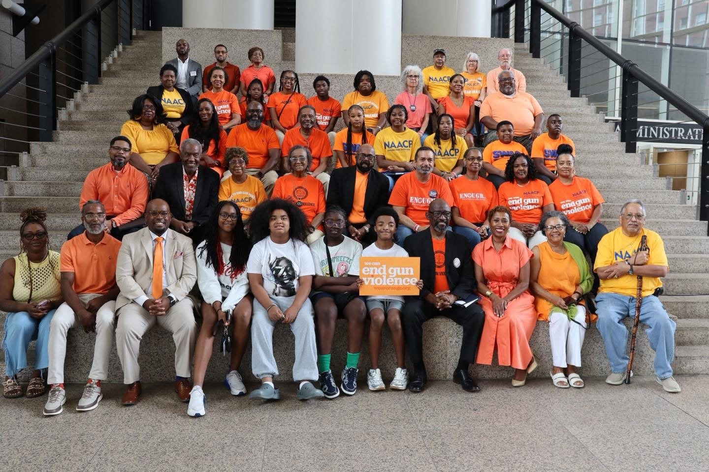 Large group photo of community leaders, residents, youth, and advocates gathered on the steps inside Mobile Government Plaza during a Project Wear Orange event. Participants are dressed primarily in orange shirts and clothing, many displaying messages supporting the effort to end gun violence. Several individuals wear NAACP apparel, while others hold signs promoting violence prevention and community safety. The diverse group poses together in a show of unity, solidarity, and commitment to raising awareness about gun violence and supporting safer communities in Mobile, Alabama.