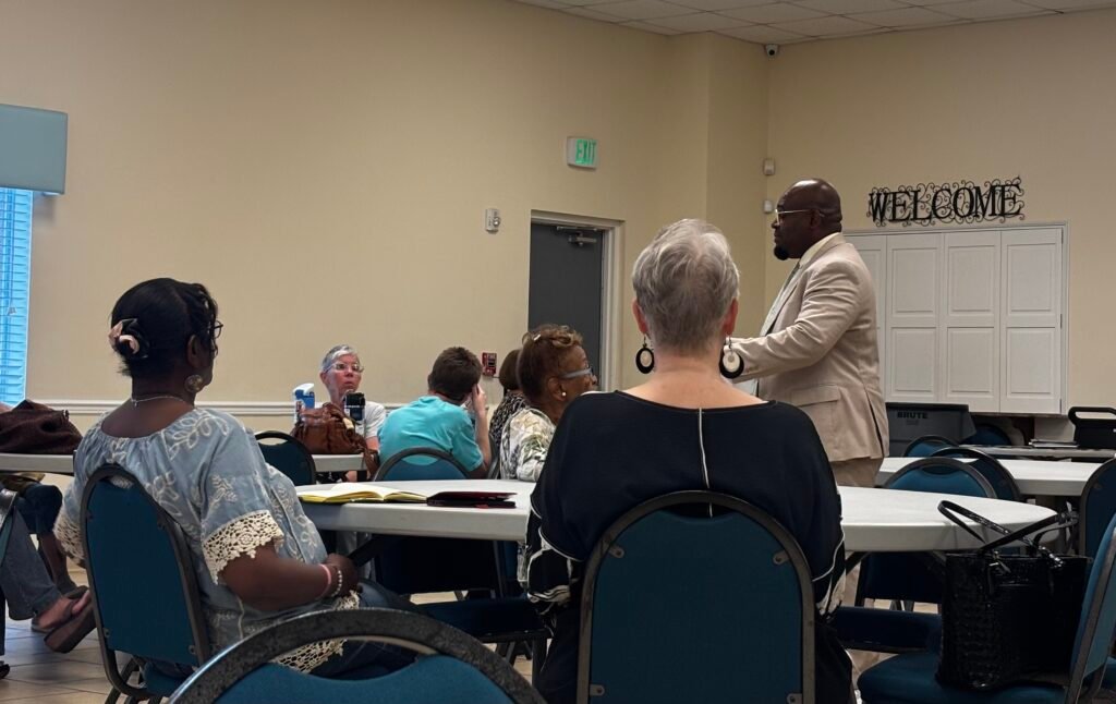 A close-up shot from behind attendees at the District 1 community meeting, showing Mobile City Councilman Cory Penn standing at a table in a tan suit jacket and glasses as he speaks and gestures with his hands.