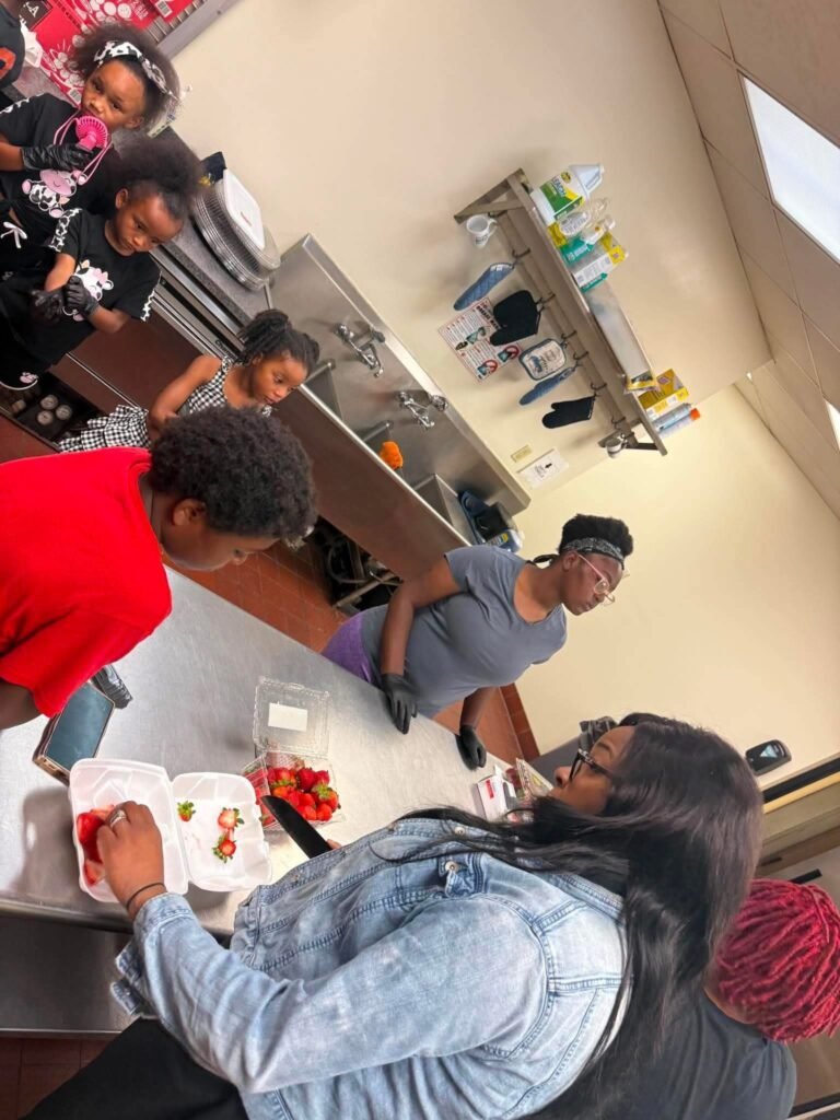 Love Speaks Inc. volunteer in a denim jacket supervises children wearing black gloves as they assemble plates of strawberries and waffles during a community cooking workshop in Mobile, Alabama.