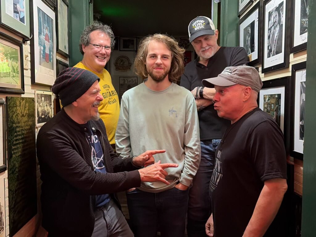A photograph featuring musicians Phil & Stan Foster alongside others standing together in a room with framed pictures on the walls.
