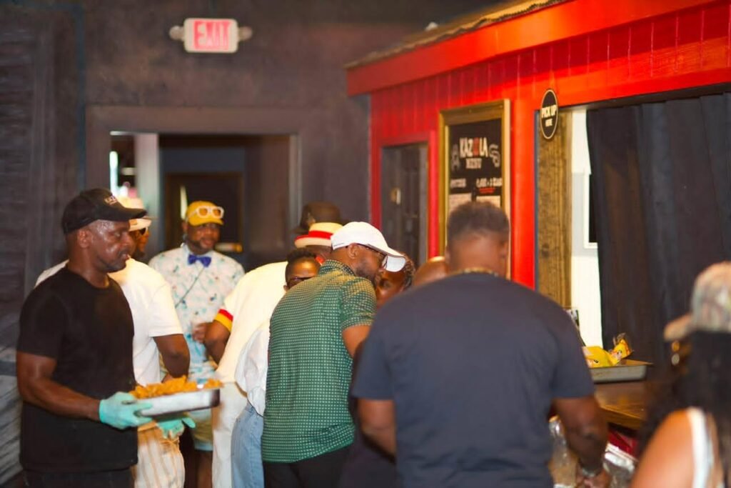 A candid shot inside Kazoola showing guests waiting at the food counter, with a staff member in a black t-shirt and gloves holding a metal tray of food in the foreground.