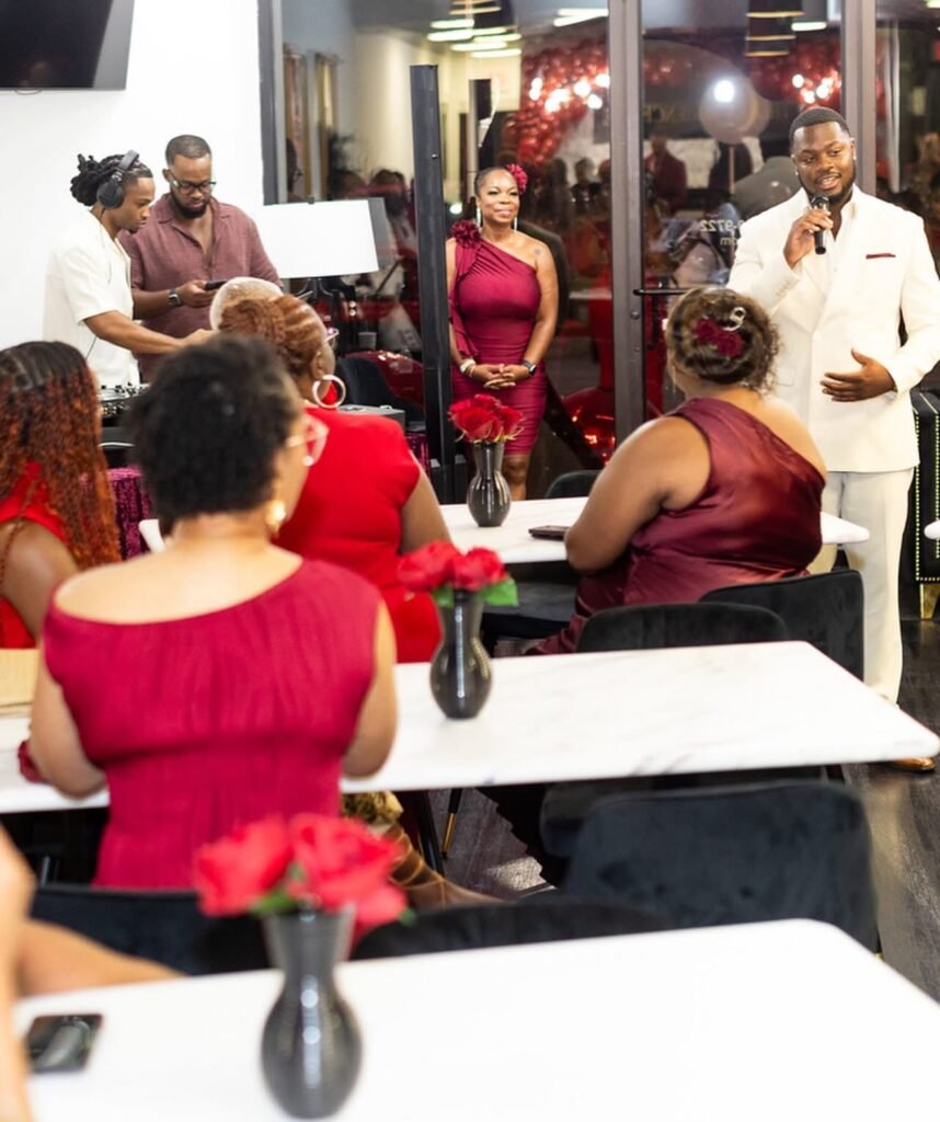 Alvin Jarreau speaking into a microphone at the front of a room during Rosé At Night, with seated guests dressed in red and burgundy facing him.