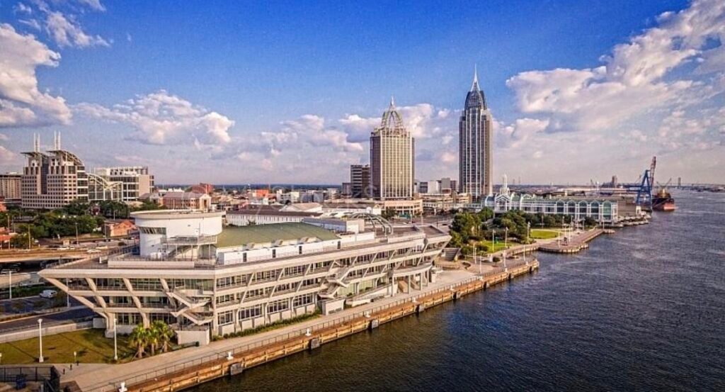 Aerial photograph showing the waterfront location of the National Maritime Museum of the Gulf in downtown Mobile, Alabama. The museum's unique ship-shaped building is prominent on the Mobile River, with the downtown skyline and RSA Tower visible in the distance under a blue sky with scattered clouds.