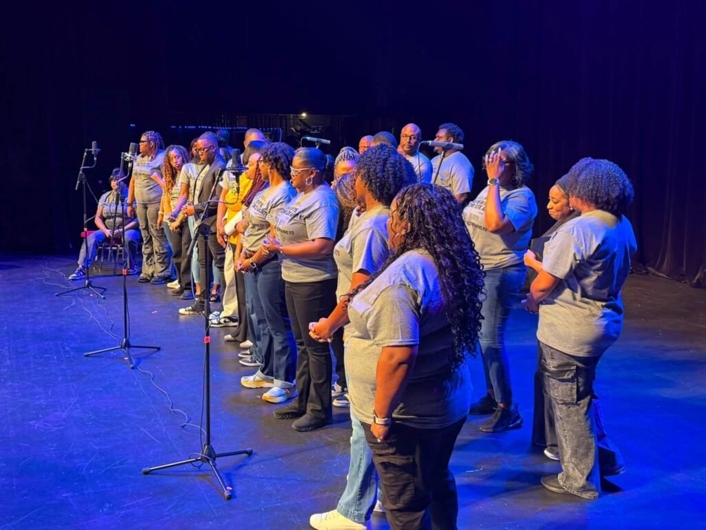 Adult choir members and participants in matching grey t-shirts standing on stage with microphones during the YOTWS reunion event.