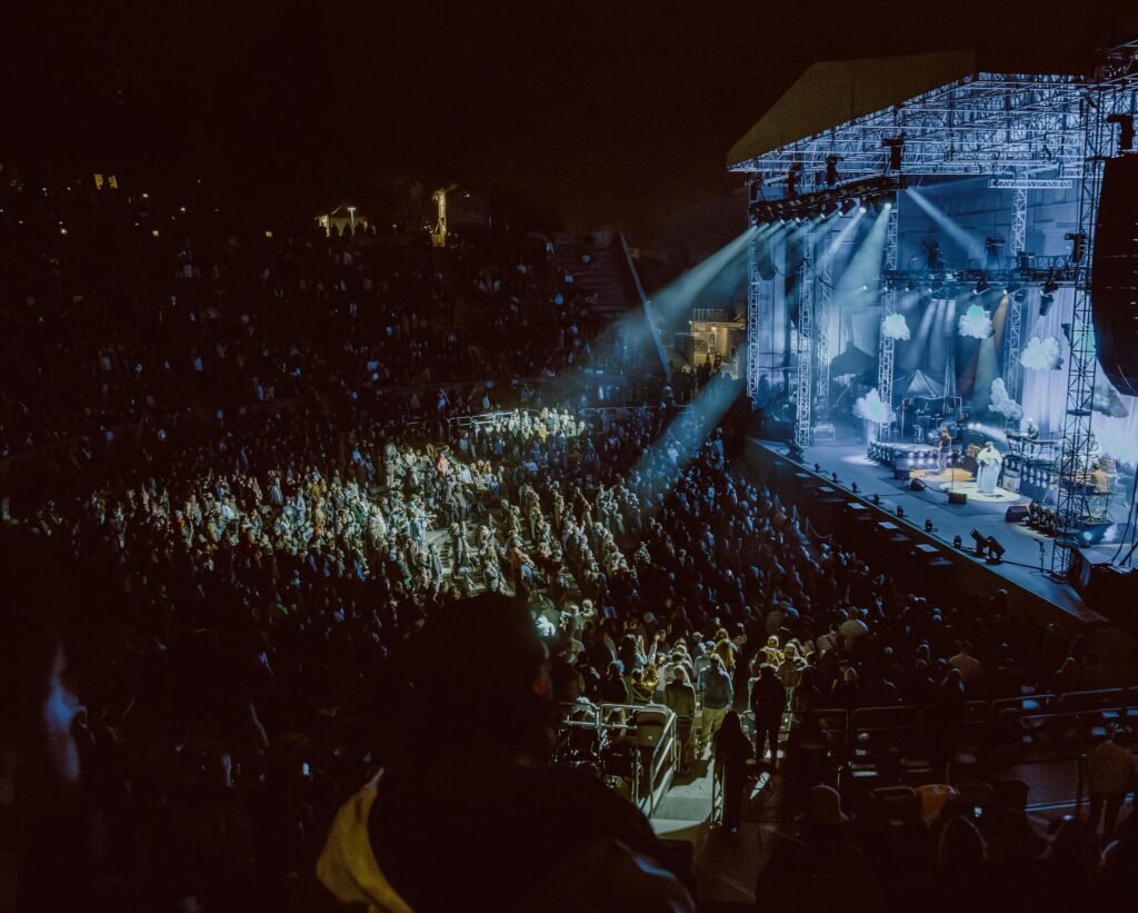 Wide shot of a nighttime concert venue featuring a large audience and Alabama Shakes performing on a brightly lit stage decorated with clouds, where they announced their new album.
