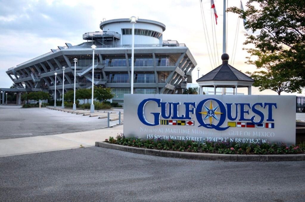 Large metallic sign at the entrance of the National Maritime Museum of the Gulf (GulfQuest) in Mobile, Alabama, featuring the museum name, address, and coordinates. The distinctive modern architecture of the museum building is visible in the background under a cloudy sky.