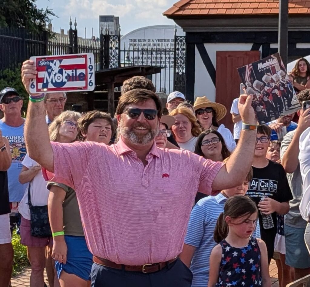 Mobile Mayor Spiro Cheriogotis, wearing sunglasses and a pink shirt, smiles while raising his arms to hold up a 1976 commemorative license plate and a historical booklet for the crowd at Colonial Fort Condé.