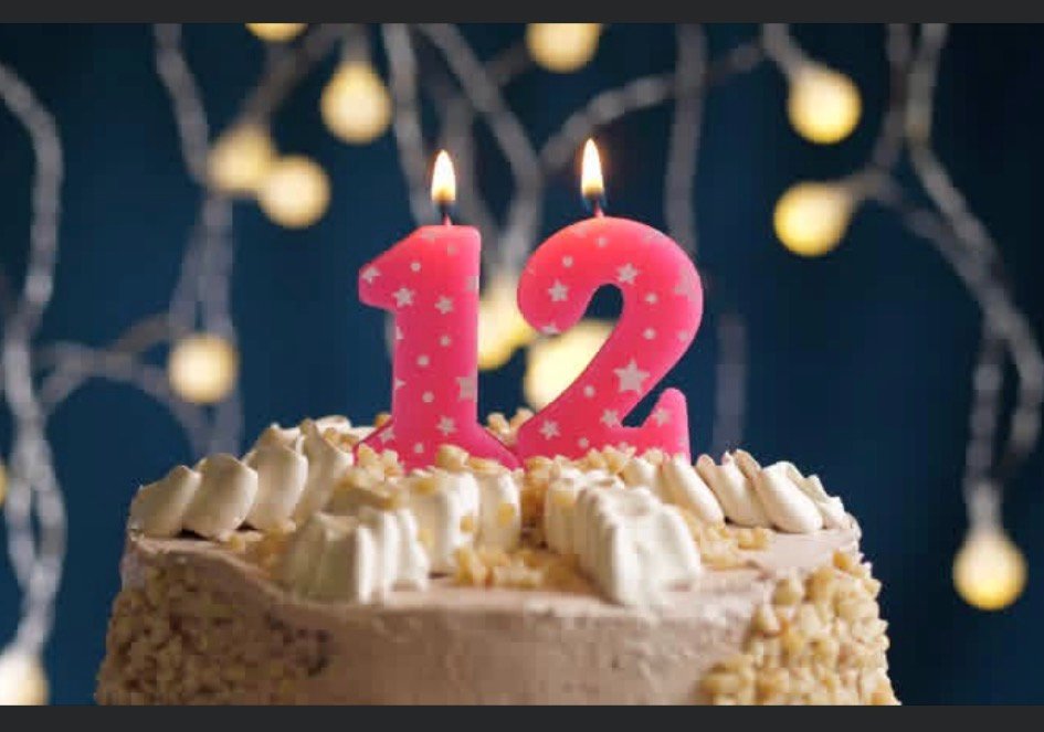 A frosted cake topped with two lit pink candles shaped like the number 12, set against a background of soft, glowing fairy lights.