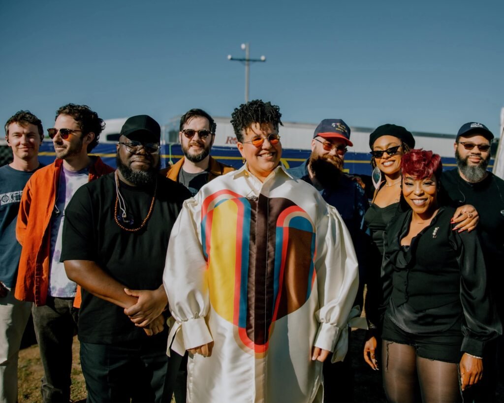 Group portrait of Alabama Shakes band members Brittany Howard, Heath Fogg, and Zac Cockrell standing together outdoors under a bright blue sky.