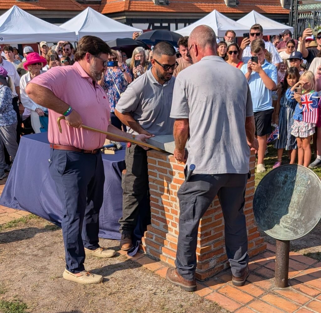 Mobile Mayor Spiro Cheriogotis, wearing a pink shirt, uses a crowbar alongside two museum staff members to pry the concrete capstone off a brick time capsule monument as a crowd watches in the background at Colonial Fort Condé.