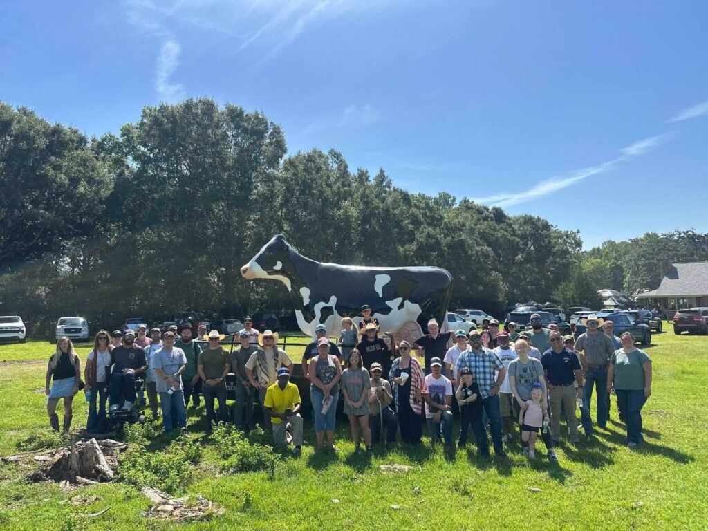 Group photo of veterans and organizers standing in front of a giant fiberglass cow statue on a sunny, grassy field at the C-4 Cattle Company in Grand Bay.