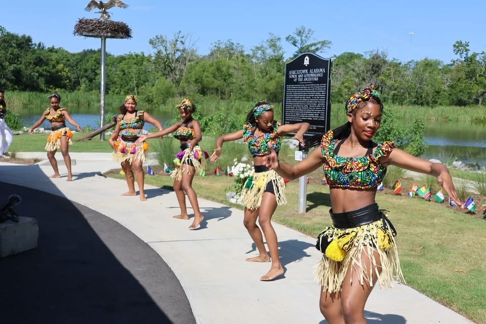 Traditional dancers in vibrant attire performing on a paved path in front of the Africatown historical marker at Lewis Landing Park, Mobile, during the Middle Passage Convening on July 10, 2026.
