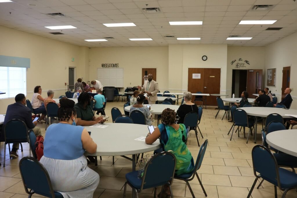 A wide shot of a brightly lit church community room with tile floors and drop ceilings. Local residents sit around circular white tables, listening to Mobile City Councilman Cory Penn speak from the center of the room.