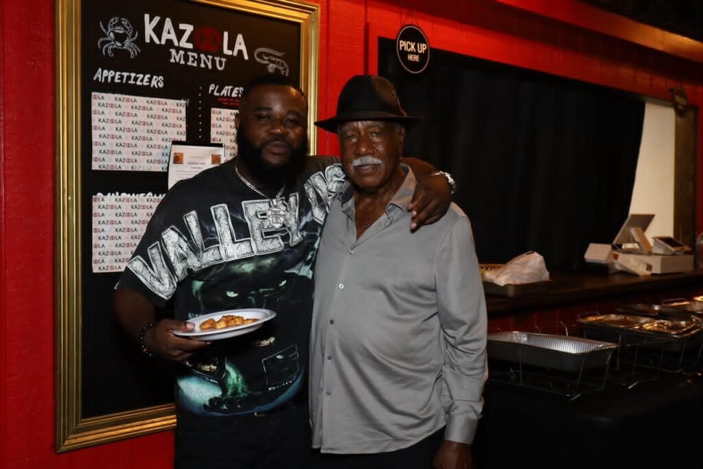 Mobile baseball legend Cleon Jones stands for a photo with a fan inside Kazoola, positioned next to the framed "Kazoola Menu" sign. Jones is wearing a black fedora, and the fan holds a small plate of food.