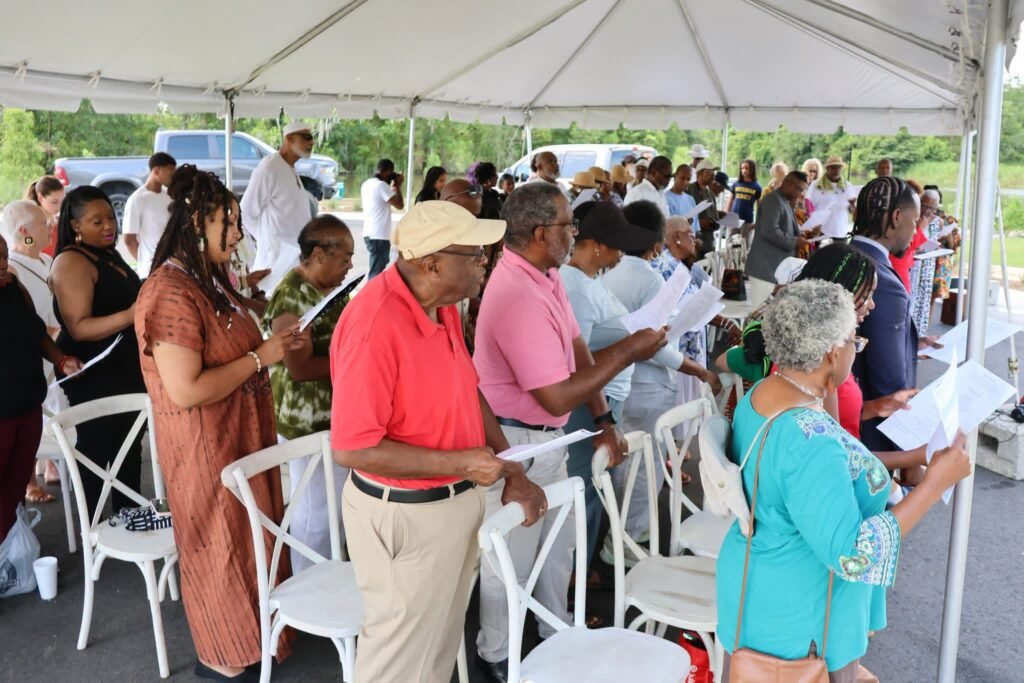 Attendees standing under an event tent at Lewis Landing Park in Mobile, Alabama, participating in a communal reading during the Africatown Middle Passage Convening on July 10, 2026.