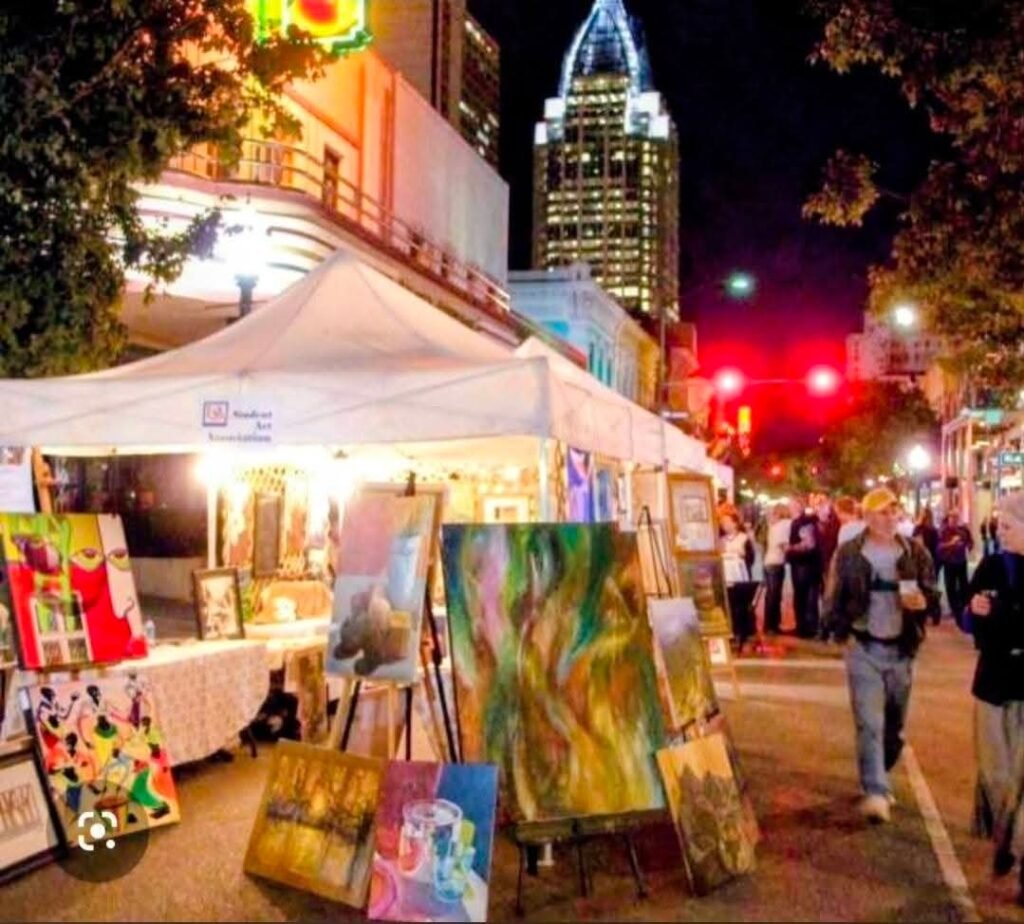 photo showing a night scene at a previous LoDa ArtWalk with artist tents, paintings on display, and visitors walking through the downtown district.