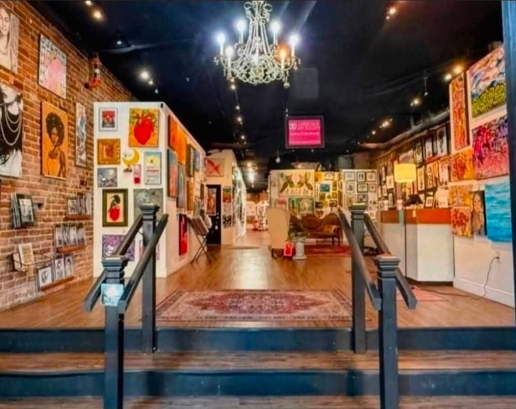 An interior view of a downtown Mobile art gallery featuring exposed brick walls, hung paintings, and a chandelier, representative of the creative venues open during the LoDa ArtWalk.