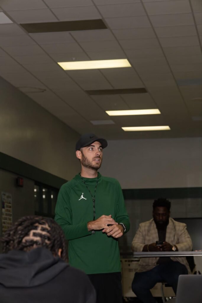 Coach Mike Hoffmann, wearing a green Jordan long-sleeve shirt and a black cap, speaks to the crowd at a Vigor High School welcome event.