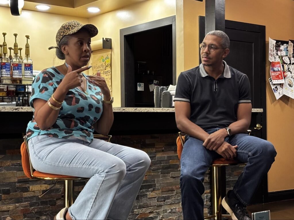 Business owner Kimberly Knight and community organizer Reggie Weaver sit on barstools inside Great Day Latte during the Civics & Conversation forum. Knight, wearing a leopard-print cap and a blue patterned shirt, gestures with her hands while speaking. Weaver sits to her right, wearing a dark polo shirt and jeans, listening attentively.