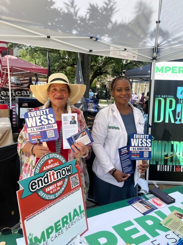 Two women smiling inside a white vendor tent at Cathedral Square, holding flyers and signs for Everett Wess alongside official endITnow sponsor and Imperial Care Services branding.