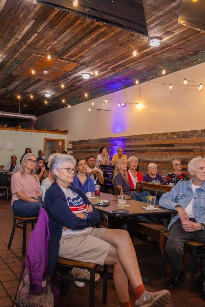 A side view of the town hall audience seated inside The Electric Piano Parlor under an overhead wooden ceiling lit with warm string lights. In the foreground, a woman wearing a blue Clyde Jones campaign t-shirt over a white long-sleeve shirt sits with her arms crossed. Other community members sit along a long wooden table and chairs stretching down the room, with a dark horizontal wood-paneled wall on the right.