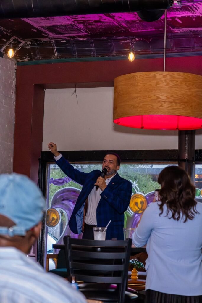 Medium shot of congressional candidate Clyde Jones in a dark suit, speaking passionately with one hand raised at a microphone during a town hall meeting.