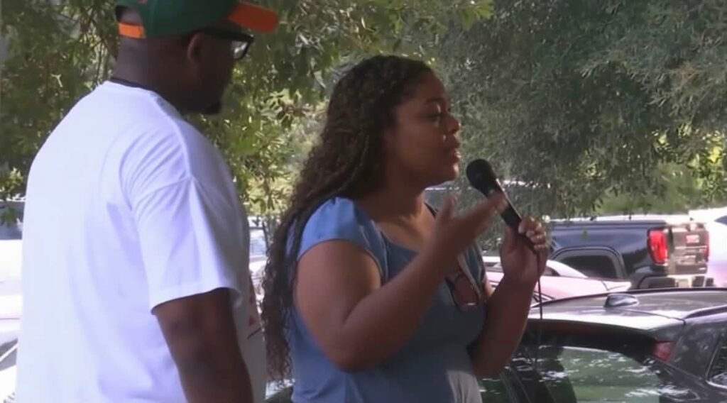 District 1 Councilman Cory Penn stands next to a woman who is speaking into a handheld microphone outdoors at Tricentennial Park.