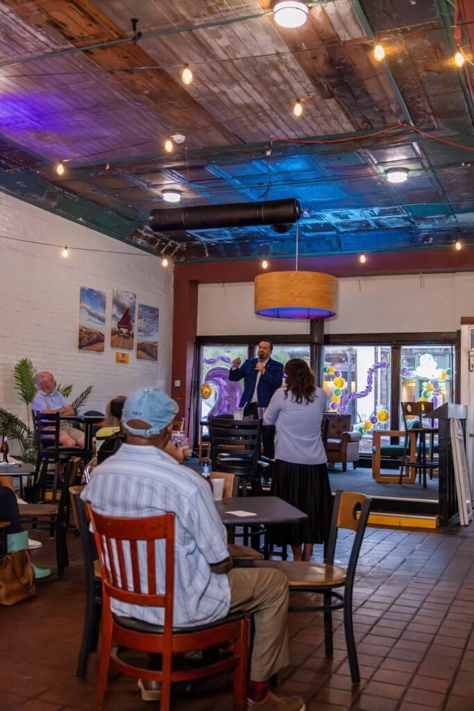 Democratic congressional candidate Clyde Jones stands at the front of The Electric Piano Parlor speaking into a microphone. He is wearing a dark blue suit jacket and a light blue dress shirt, gesturing with his hands as an organizer stands next to him. An audience sits at small black tables in the foreground under hanging warm string lights, while a wall on the left features three framed landscape photos of a red piano. Large glass doors and colorful balloon decorations are visible at the back entrance.