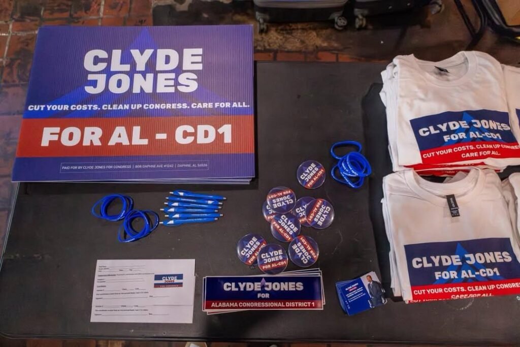 A dark tabletop displays a collection of Clyde Jones campaign merchandise for Alabama's 1st Congressional District (AL-CD1). The items include a large stack of blue and red signs reading "Clyde Jones For AL - CD1," folded white t-shirts with matching campaign branding, stacks of blue ink pens, round campaign buttons, blue wristbands, signup sheets, and business cards featuring a portrait of the candidate.