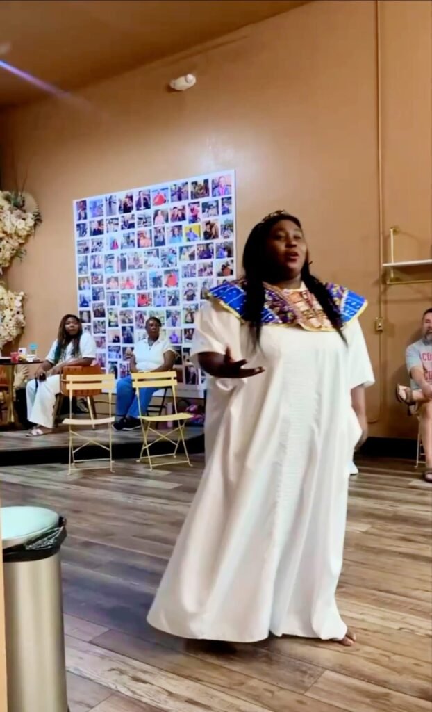 Ashley Waller, performing as Queen Black, stands in the center of Great Day Latte wearing a long white kaftan robe with a blue and gold pattern around the collar. She has her hand extended while performing for a seated audience, with a large collage of small photos hanging on the wall behind her.