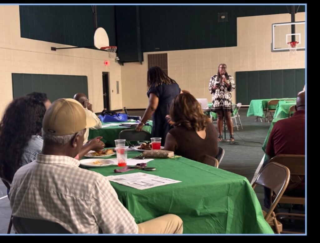 Shonnda Smith stands at a microphone at the front of a gymnasium-style room, wearing a black and white patterned dress. An audience sits in folding chairs at a long table covered in a green tablecloth in the foreground, with plates of food and cups visible on the table. A basketball hoop hangs on the wall in the upper right background.