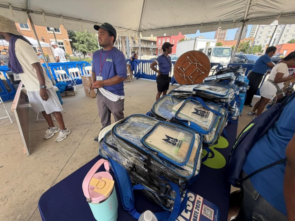 Stacks of clear plastic backpacks filled with school supplies rest on a blue table under a white event tent. The notebooks inside the backpacks display the Mobile Parks and Recreation Department logo. A staff member in a blue department shirt stands behind the table, with other workers and event equipment visible in the background.