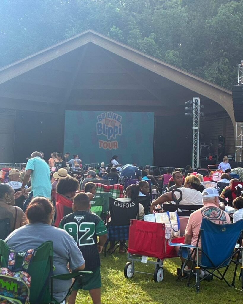 Families and children gathered on lawn chairs at Medal of Honor Park, facing the pavilion stage where the "Be Like Blippi Tour" screen is displayed.