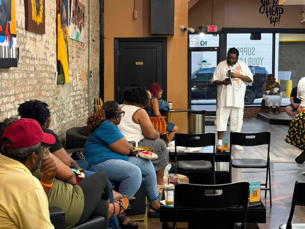 Alex Jerome Lofton stands at the front of Great Day Latte cafe wearing an all-white shorts set. He holds a microphone and looks down at his phone while an audience sits in chairs and on barstools watching him. The cafe features brick walls with artwork on the left and a glass entrance doors in the background.