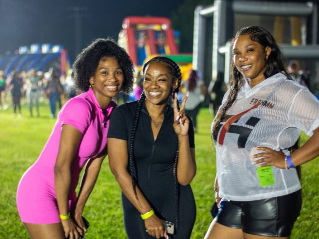 Three women smiling together outdoors at night during the Inflatable Night Grown Up Edition at Medal of Honor Park, with illuminated inflatables visible in the background.