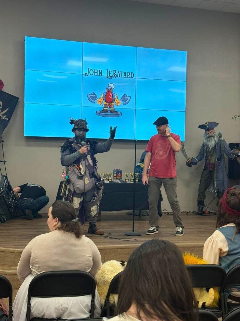 John LeBatard Sr., dressed in an elaborate fantasy steampunk outfit, speaks into a microphone next to co-host Nathan Leccese on the wooden stage at the Semmes Community Center. The screen behind them displays a cartoon viking graphic titled "John LeBatard," with a life-sized pirate prop standing nearby on the stage floor.