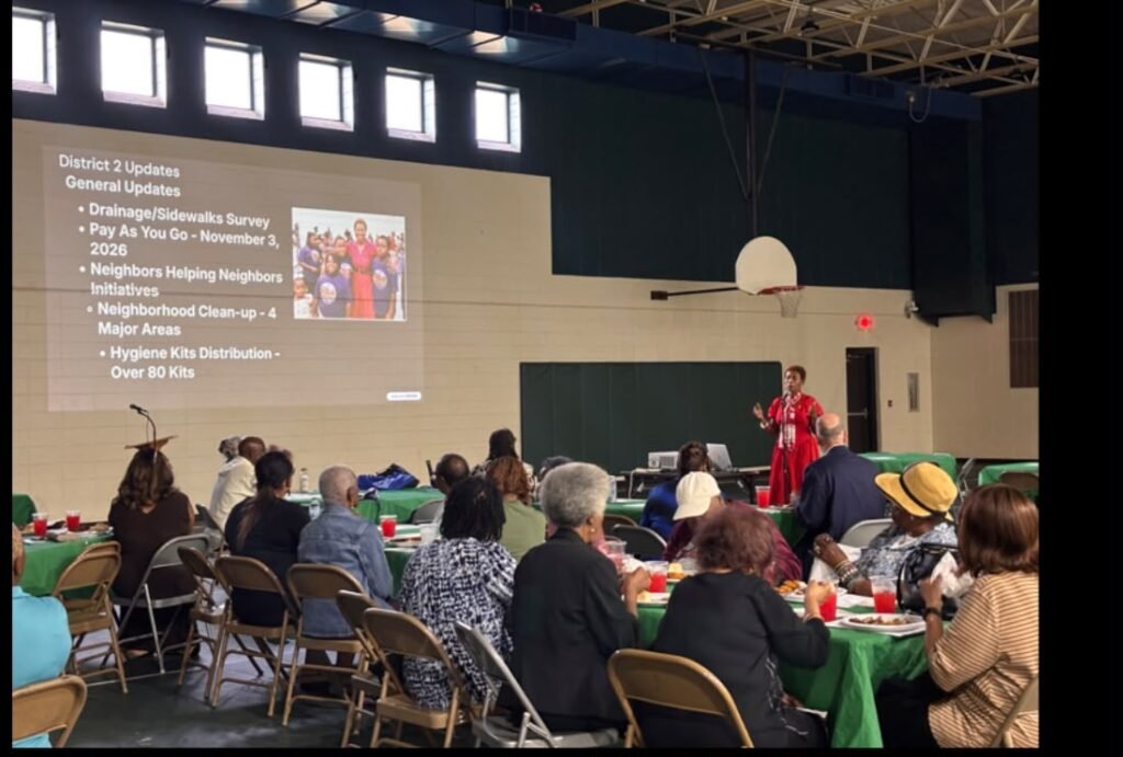 Councilwoman Samantha Ingram stands at the front of a gymnasium-style room speaking into a microphone. She is wearing a red outfit and gesturing with both hands. To her left, a large projector screen displays a slide titled "District 2 Updates General Updates," listing bullet points for the Drainage and Sidewalks Survey, the November 3 Pay As You Go referendum, and the Neighbors Helping Neighbors Initiatives alongside a small photo. An audience sits at circular tables with green cloths watching her presentation.