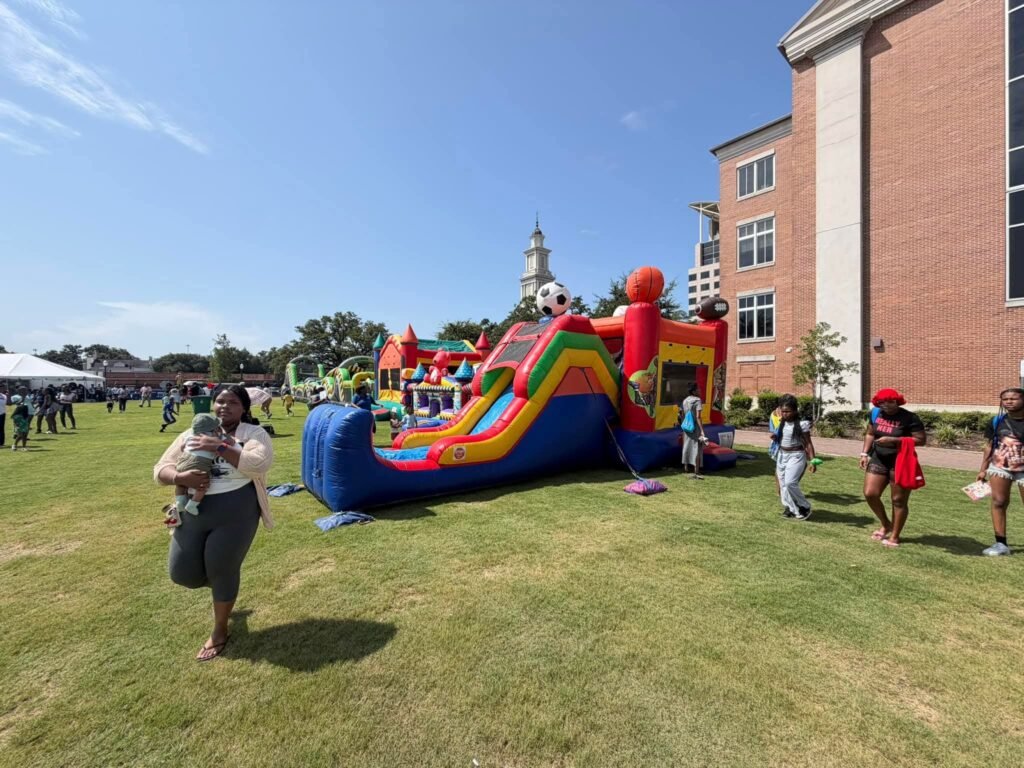A bright, sunny outdoor scene at Mardi Gras Park featuring large colorful inflatable bounce houses and slides set up on a green grass field. In the foreground, a woman walks while carrying a young child, while other families and children are seen walking across the lawn nearby. A brick municipal building stands on the right side under a clear blue sky.