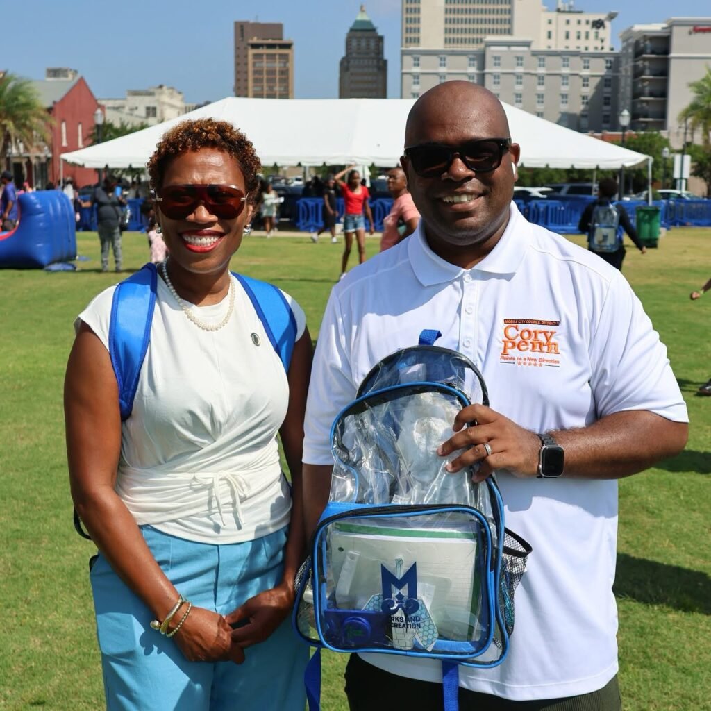 Councilwoman Samantha Ingram and Councilman Cory Penn stand side-by-side smiling on the lawn at Mardi Gras Park. Councilwoman Ingram is on the left wearing sunglasses, a white shirt, blue pants, and a blue backpack. Councilman Penn stands on the right wearing sunglasses and a white polo shirt featuring his campaign logo. He holds up a clear plastic backpack packed with blue and green school supplies displaying the Mobile Parks and Recreation Department logo. A large white event tent and city buildings are visible in the background.