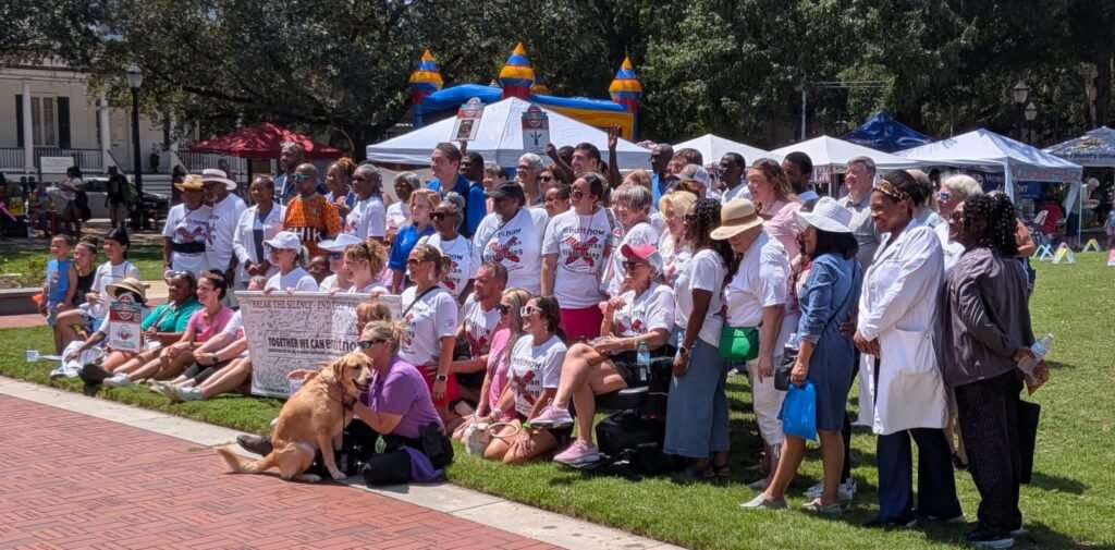 A large group of diverse community members, organizers, and a service dog gathered outdoors on the lawn at Cathedral Square for a group photo during the event.