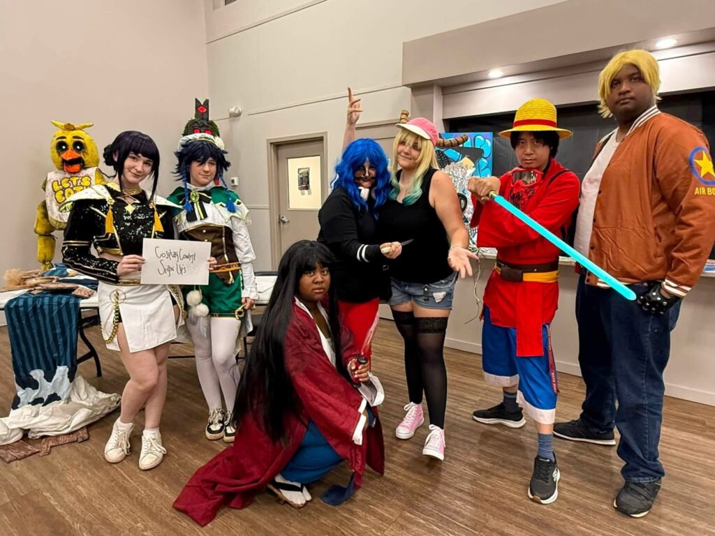 A diverse group of seven cosplayers poses together inside the Semmes Community Center next to a table with a sign that reads "Cosplay Contest Sign Ups." The characters represented include Chica from Five Nights at Freddy's, Venti from Genshin Impact, Monkey D. Luffy from One Piece, and other anime and video game characters holding props like a glowing blue sword.