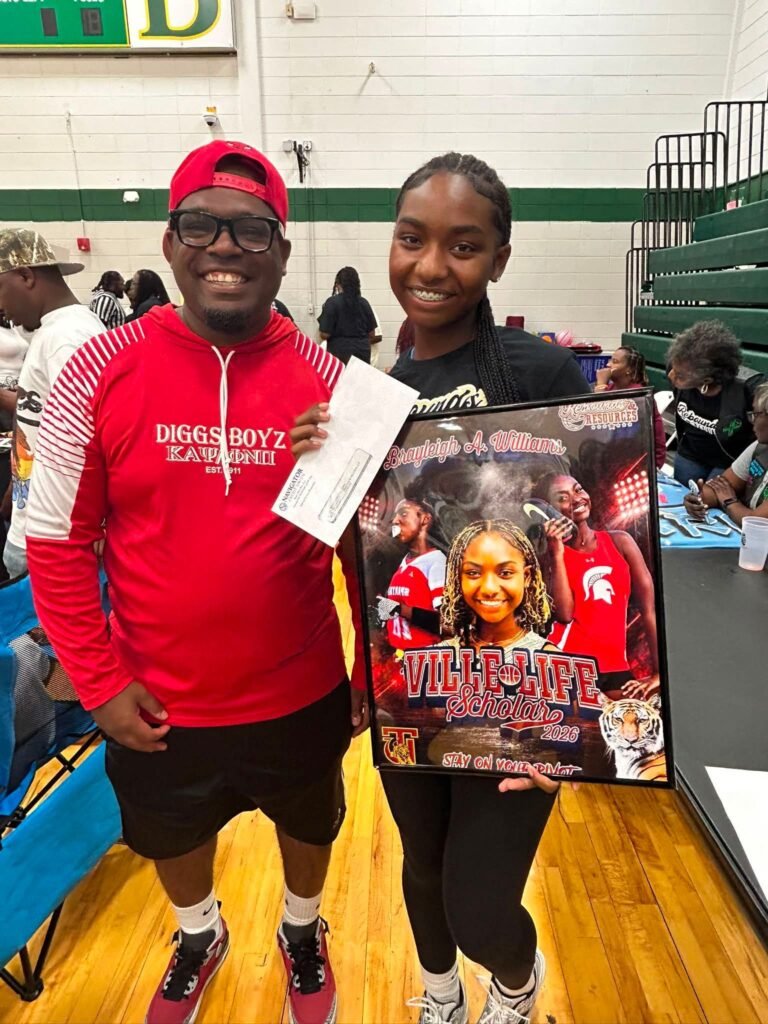 City Councilman Cory Penn and a young woman holding her "Ville Life Scholar 2026" award poster inside the Bishop State Community College gymnasium.