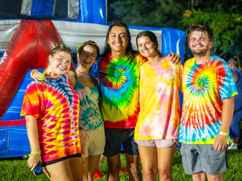A group of five smiling adults wearing colorful tie-dye shirts pose together at night in front of large inflatable structures at Medal of Honor Park.