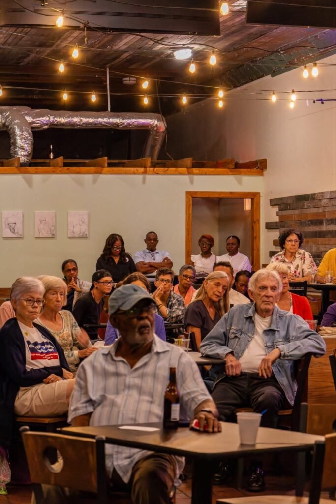 An audience of local residents sits at tables and chairs inside the dimly lit Electric Piano Parlor under warm string lights hanging across the ceiling. A man in a light blue short-sleeve shirt and cap sits in the foreground at a table, while other community members look toward the front of the room. The venue features light green walls decorated with framed artwork on the left and a wood-paneled wall on the right.