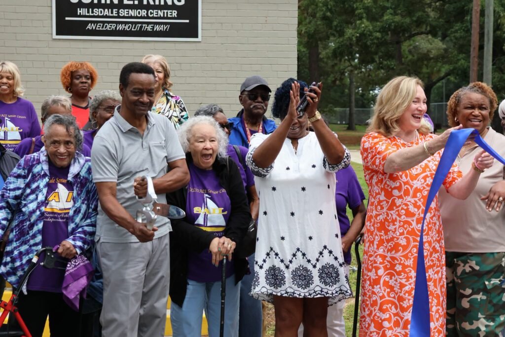 Close-up shot of local seniors, community members, and Council Vice President Gina Gregory smiling and cheering as a blue ribbon is cut in front of the John E. King Hillsdale Senior Center sign.