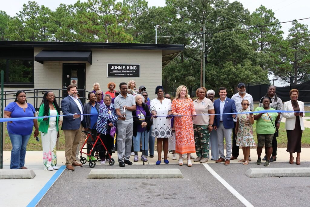 Local community members, seniors, and city officials holding a blue ribbon across the entrance of the John E. King Hillsdale Senior Center during a grand opening ceremony.